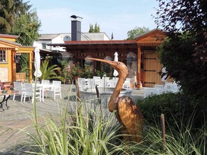 En utomhusplats med en uteplats, vita stolar och bord, en träpergola och en stor fågelskulptur.