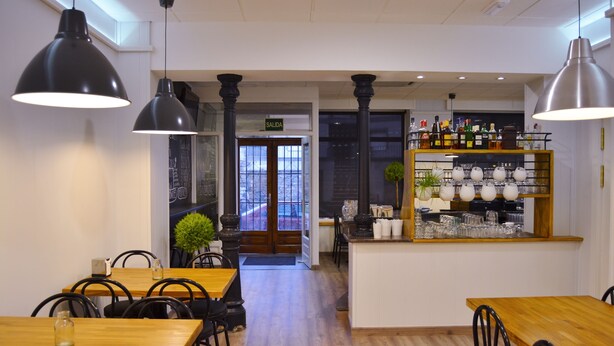 L’intérieur d’un restaurant avec des tables en bois, des chaises noires, des luminaires suspendus, un bar avec des verres et des bouteilles, et une vue à travers une porte vitrée.