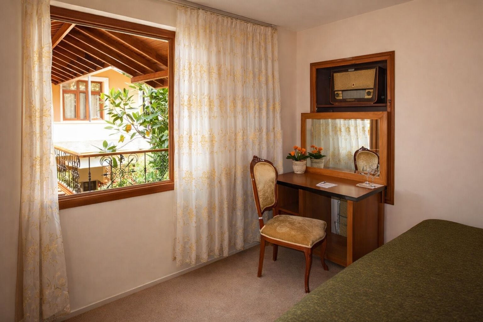 Un dormitorio con un escritorio de madera, una silla y una cama. Hay una ventana con cortinas y vistas al verdor exterior.