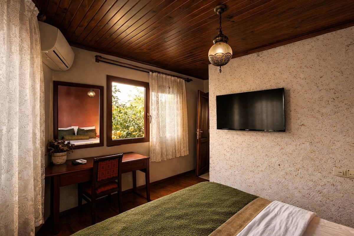 A bedroom with a wooden ceiling, a desk with a chair, a bed with green and white bedding, a TV mounted on the wall, and a window with curtains.