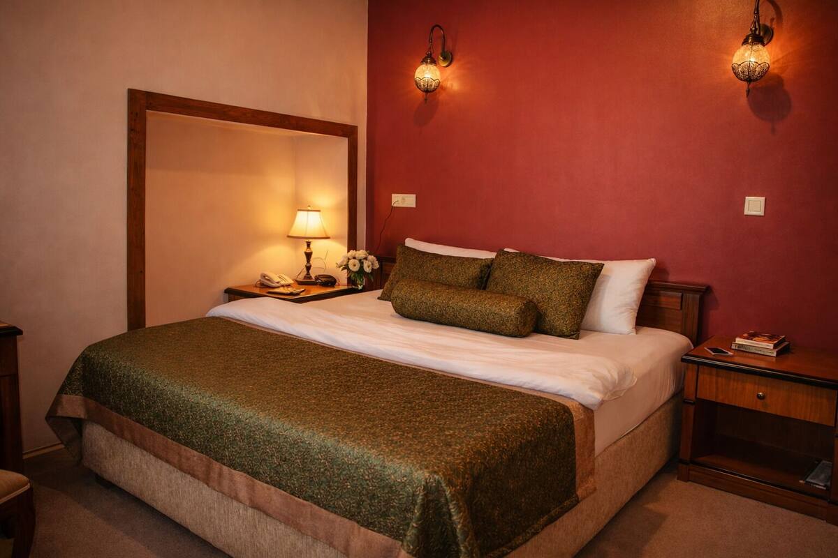 A neatly made bed with a green blanket, a bedside lamp, and a nightstand with a phone and a book.
