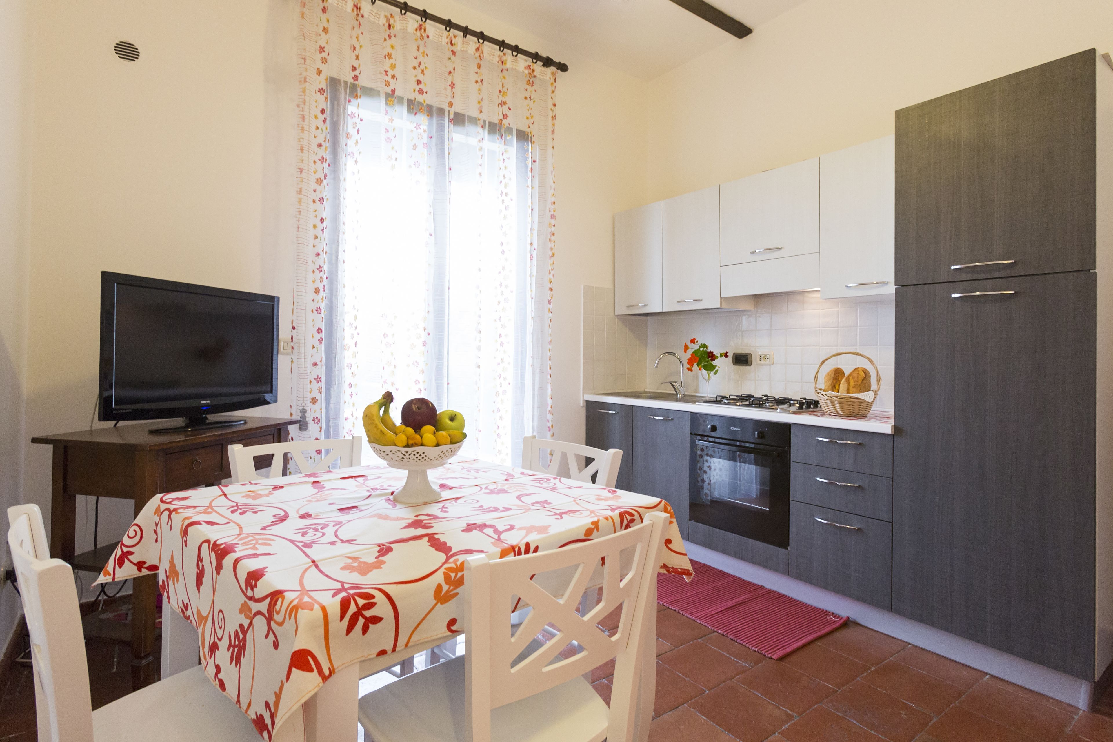 Cocina pequeña con gabinetes blancos, estufa y lavamanos. Hay una mesa de comedor con un frutero y un televisor sobre un mueble.