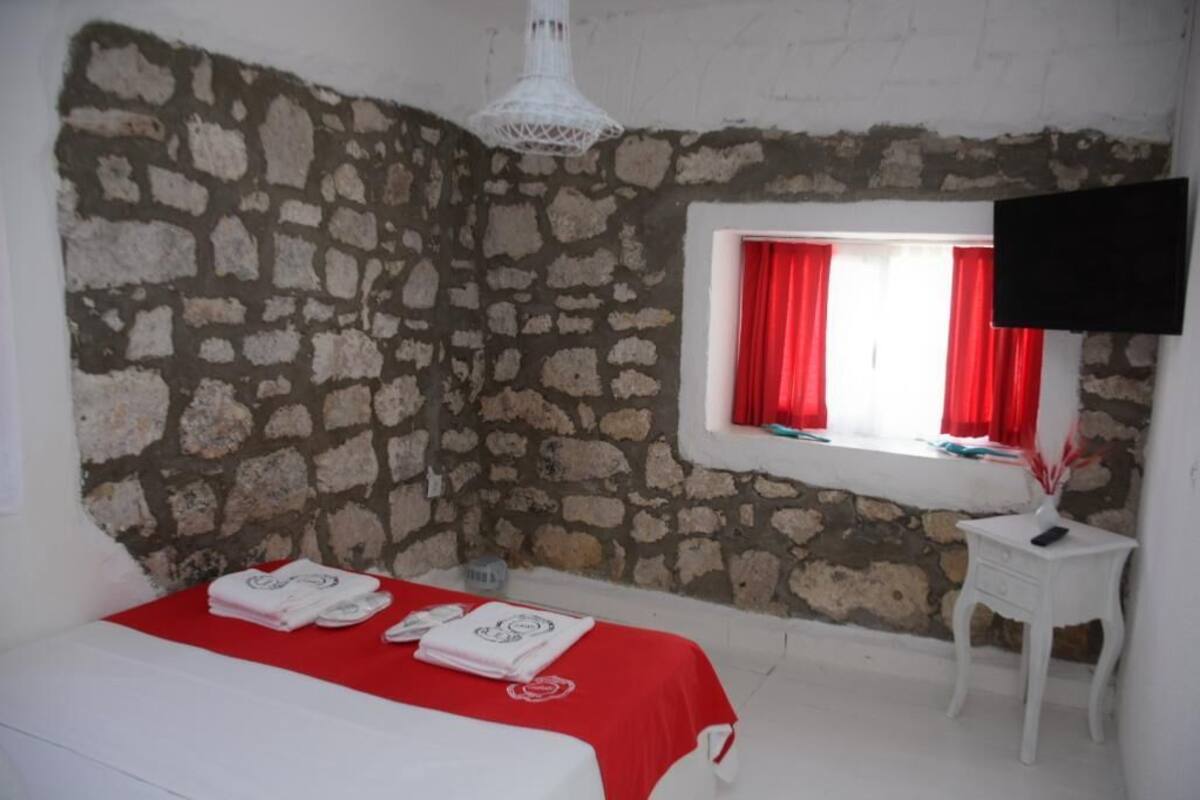 A room with a stone wall, a bed with a red bedspread, a small white table, and a window with red curtains.
