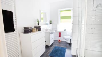 Un baño moderno con muebles blancos, un lavamanos y un inodoro. Hay toallas apiladas en la encimera y una alfombra azul en el suelo.