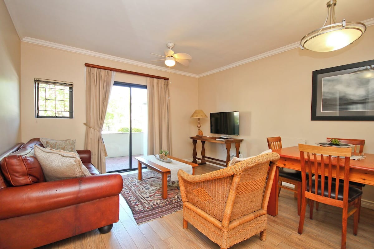 A living room with a sofa, a wicker chair, a dining table with chairs, a television, and a ceiling fan.