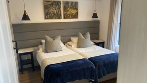 Una cama doble con cabecera gris, dos mesitas de noche y un tocador. Se observan dos unidades de aire acondicionado fijadas a la pared y dos lámparas colgantes.