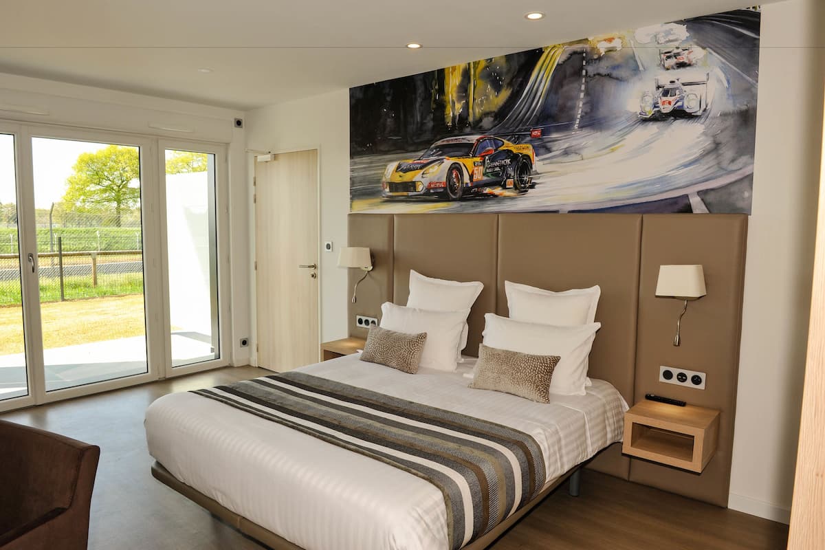 A modern hotel room with a large bed, a painting of a racing car, and a sliding glass door to the outside.