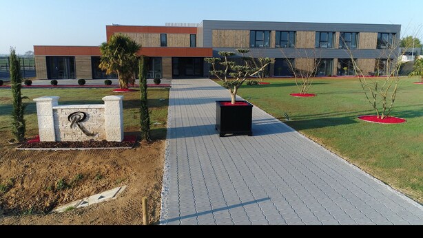 A modern building with a paved walkway, potted plants, and a stone pillar with a logo. - Résidence des Pilotes (Le Vigeant)