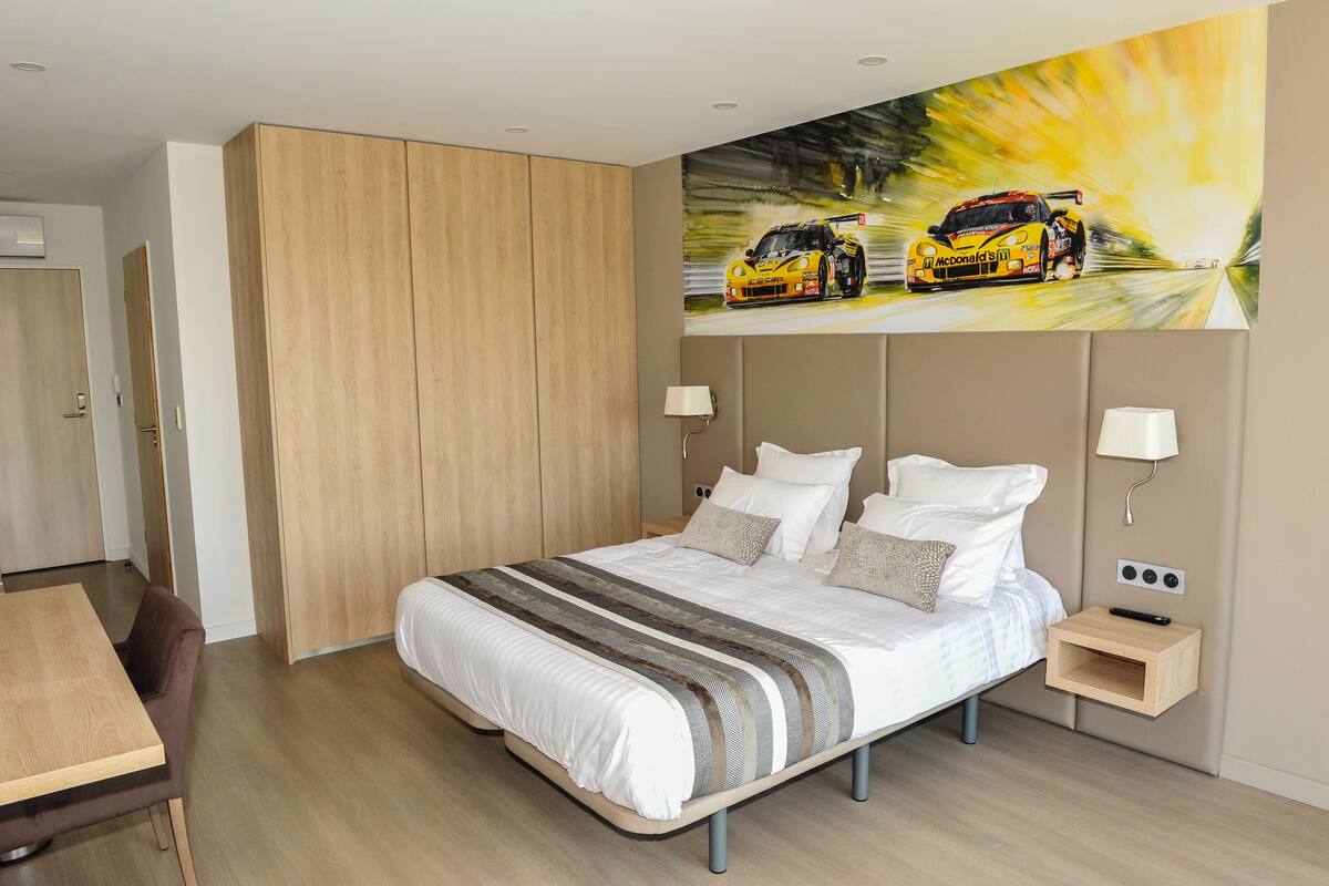 A modern hotel room with a large bed, bedside tables, a desk, and a painting of a racing car on the wall.