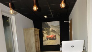 A reception desk with a vintage design, a laptop on top, and a painting of a car on the wall behind. - Résidence des Pilotes (Le Vigeant)