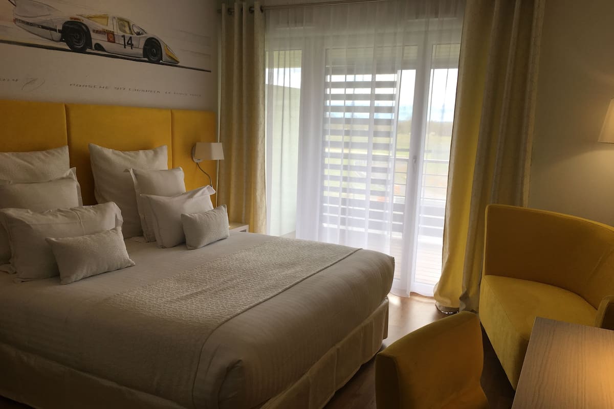 A hotel room with a large bed, a headboard with a poster, a window with curtains, and a yellow armchair.