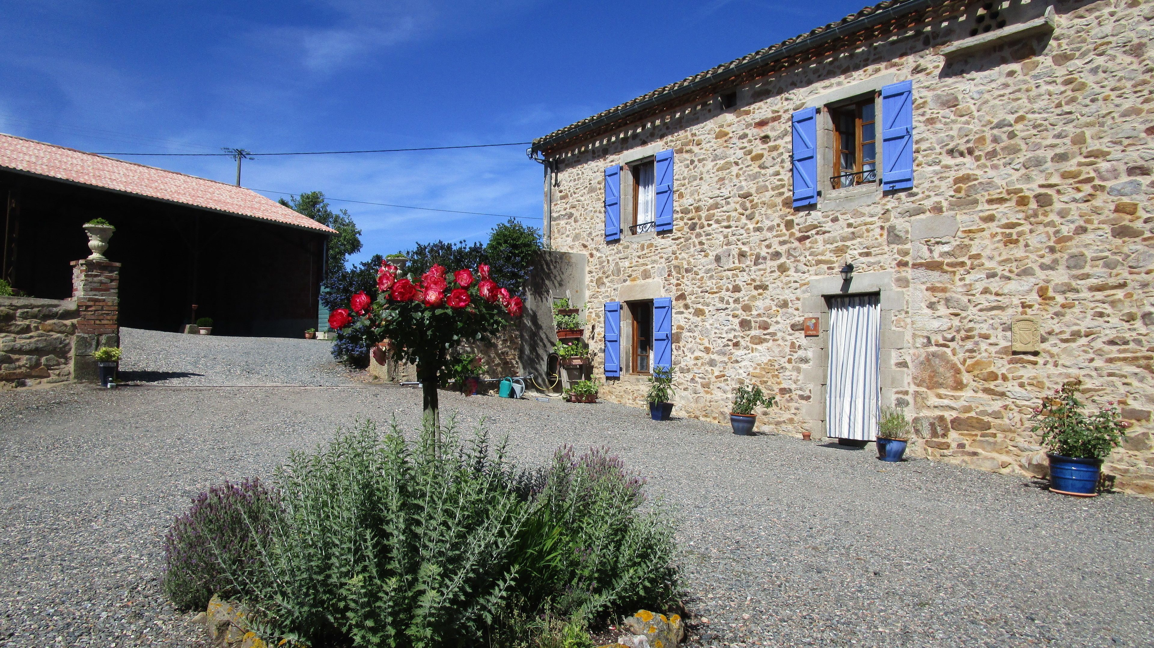 Un edificio de piedra con contraventanas azules, un patio de grava y macetas con plantas.