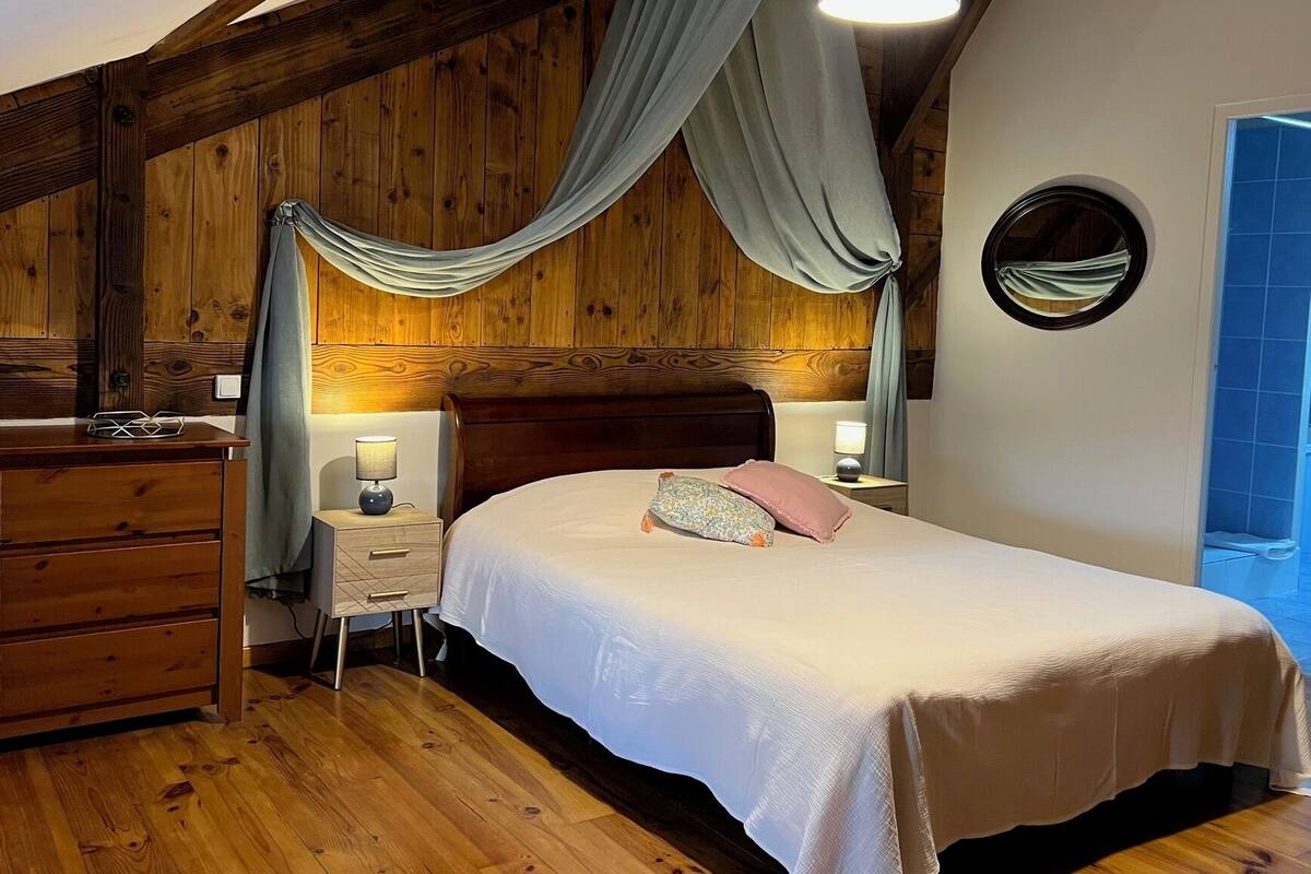 A bedroom with a wooden ceiling, a bed with a white bedspread, two bedside lamps, a wooden dresser, and a window with blue curtains.