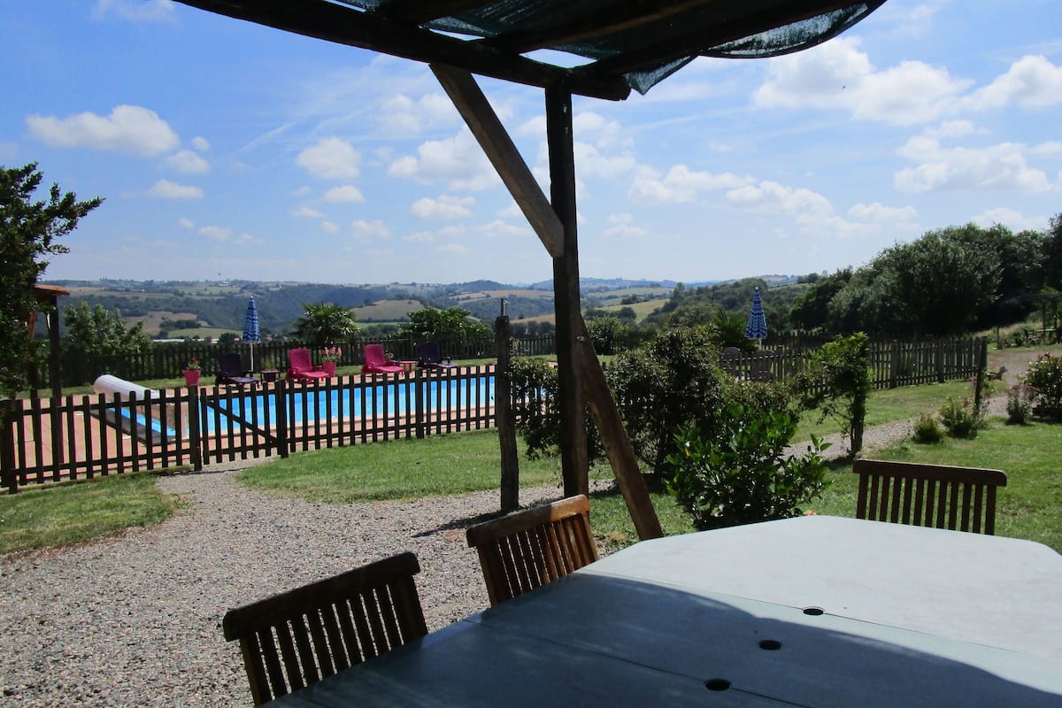 A covered outdoor seating area with a table and chairs, a pool, and a view of a hilly landscape.