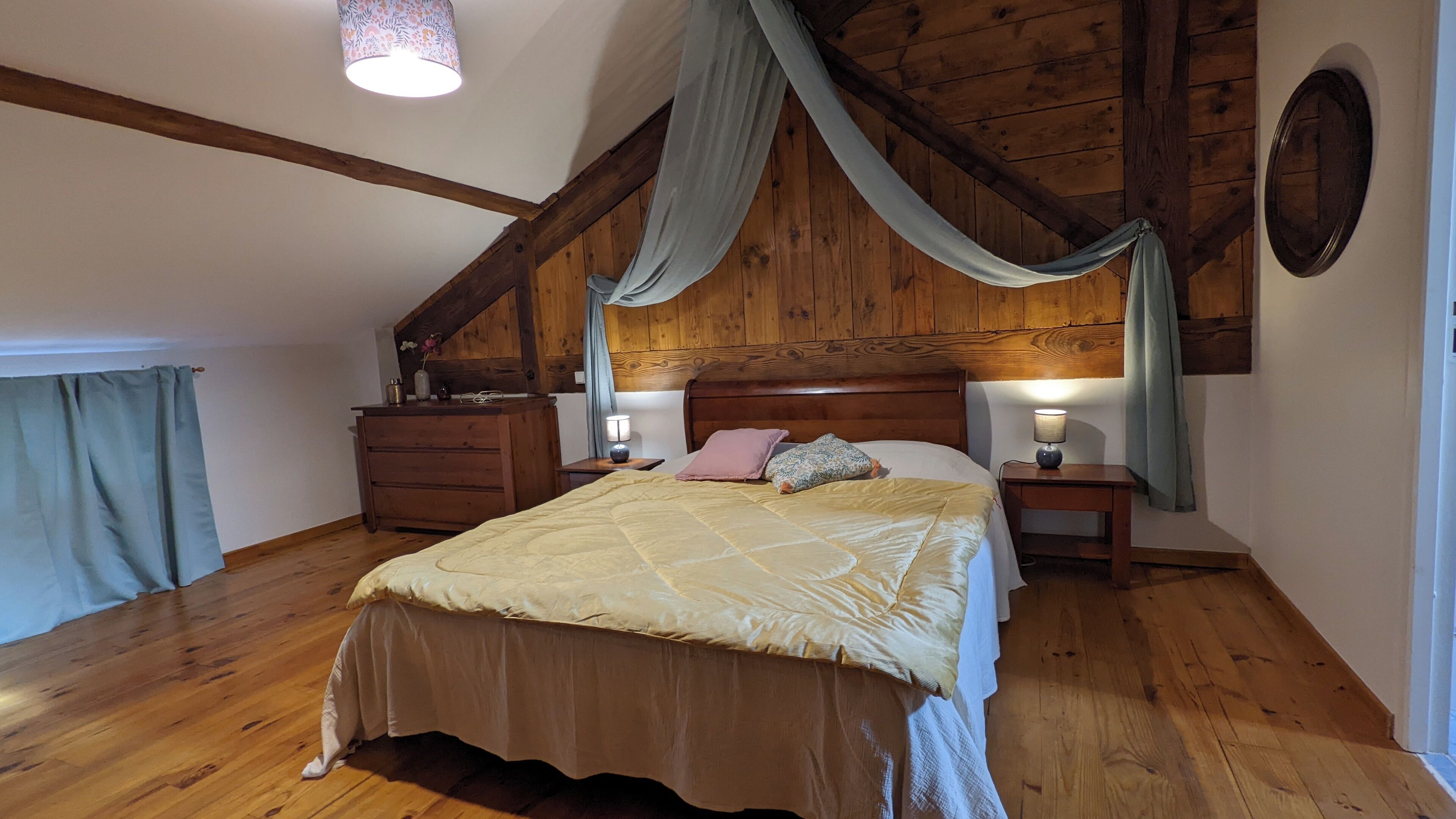 A bedroom with a wooden ceiling, a bed with a quilt, two bedside tables with lamps, a dresser, and a window with curtains.