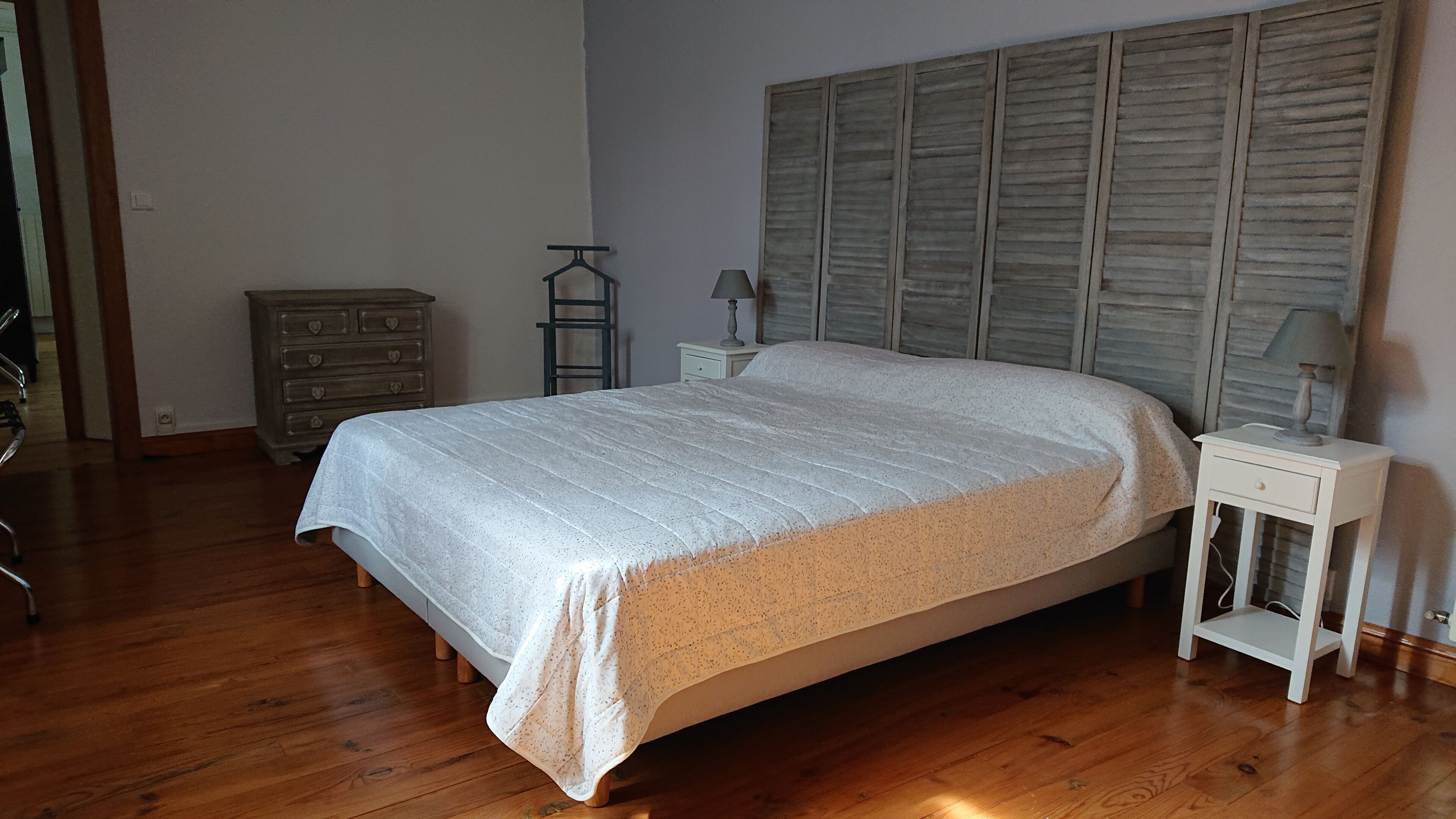 A bedroom with a wooden floor, a bed with a white quilt, a nightstand with a lamp, and a dresser with heart-shaped handles.