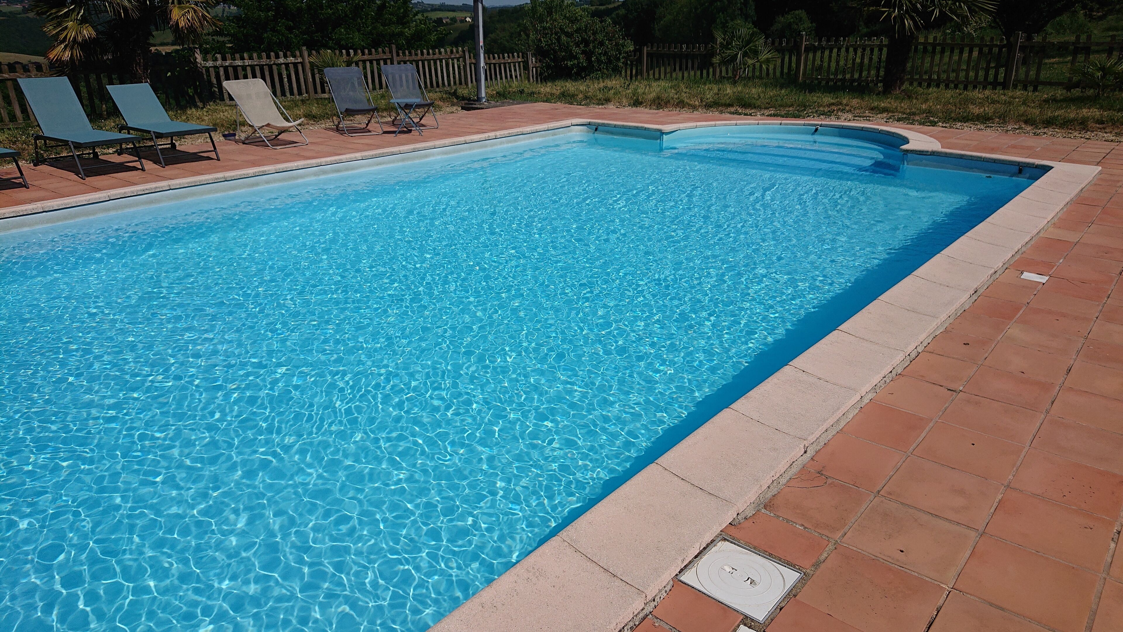 A rectangular swimming pool with clear blue water, surrounded by a tiled deck and a wooden fence.