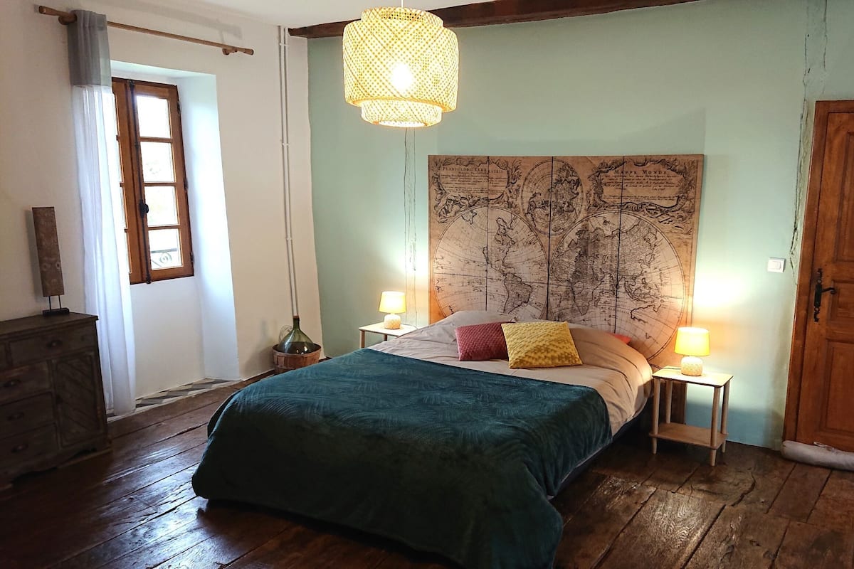 A bedroom with a large bed, wooden floor, a world map on the wall, a wooden door, and a window with curtains.