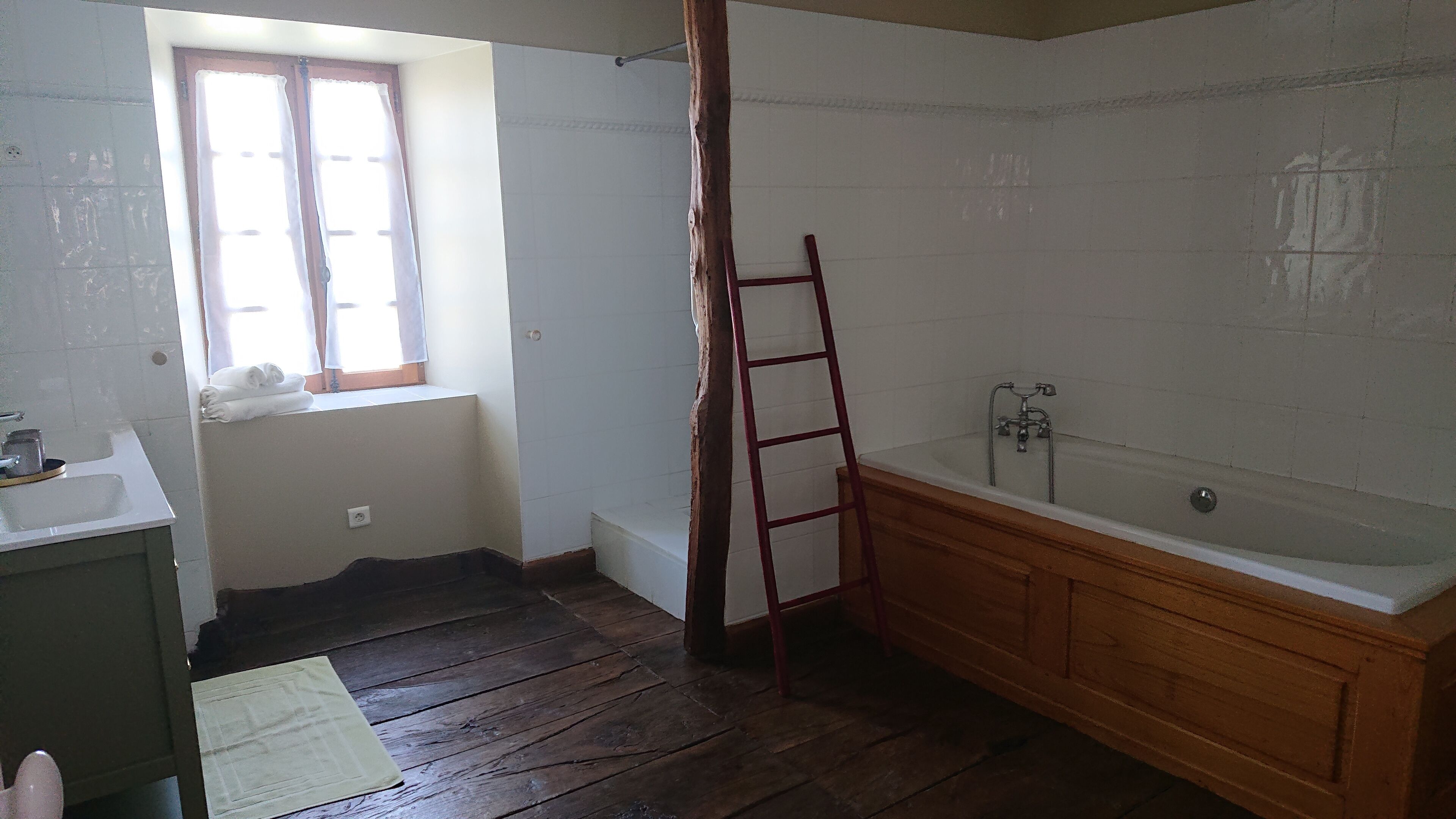 A bathroom with a wooden floor, a wooden bathtub, a window with curtains, and a white sink.