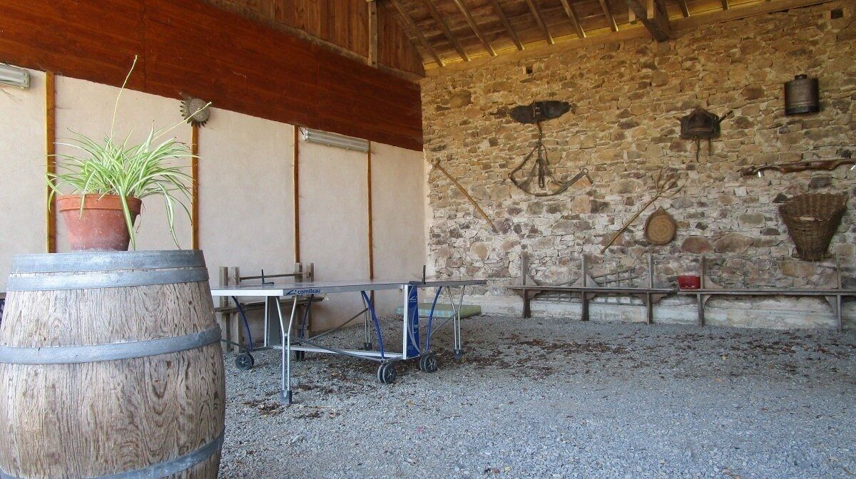 A rustic barn interior with a wooden barrel, a metal cart, and a stone wall with various tools and baskets.