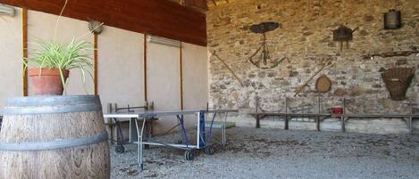 A rustic barn interior with a wooden barrel, a metal cart, and a stone wall with various tools and baskets.
