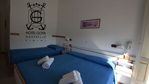 A hotel room with two beds, each with a blue bedspread and rolled towels. A logo for Hotel Goya Marebello Rimini is visible on the wall. - Hotel Goya (Rimini)