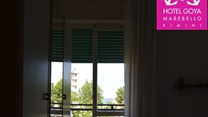 A hotel room with a bed, a bench, a wardrobe, and a balcony with a view of greenery and a building. - Hotel Goya (Rimini)