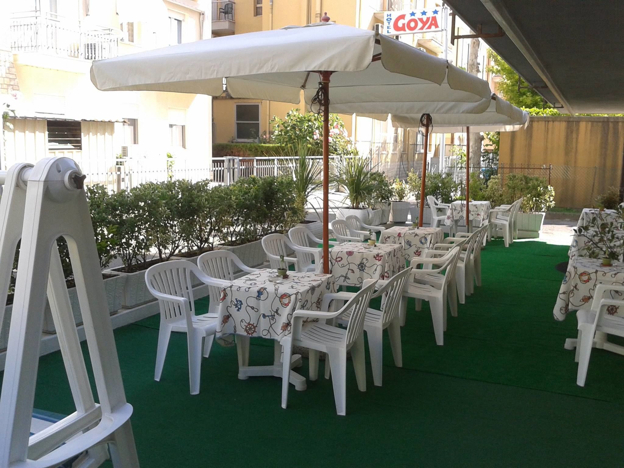 Un patio al aire libre con sillas y mesas de plástico blanco, cubierto por grandes sombrillas, y un piso de césped artificial verde.