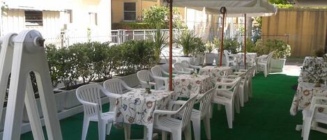 Un patio al aire libre con sillas y mesas de plástico blanco, cubierto por grandes sombrillas, y un piso de césped artificial verde.