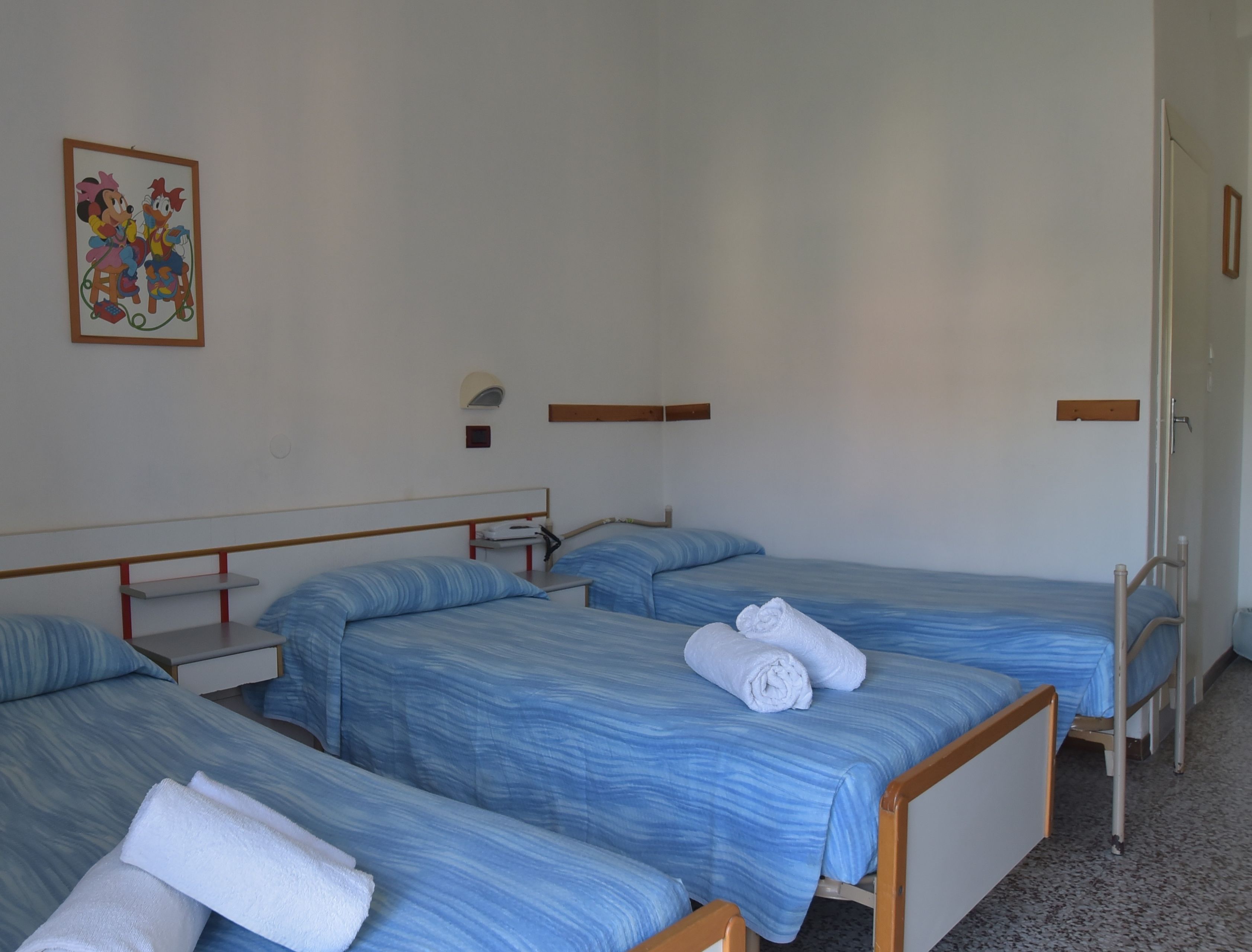 A hotel room with three single beds, each with a blue striped bedspread and white towels. There is a small bedside table with a lamp and a framed picture on the wall above the beds.