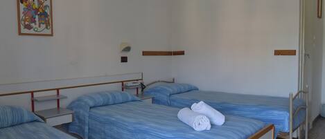 A hotel room with three single beds, each with a blue striped bedspread and white towels. There is a small bedside table with a lamp and a framed picture on the wall above the beds.