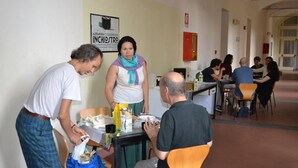Eine Gruppe von Personen in einem Raum mit Tisch und Stühlen, einem Poster an der Wand und einem Korridor im Hintergrund.