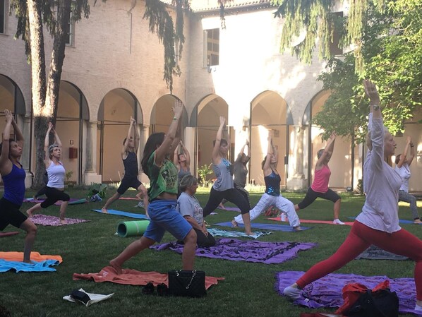 Un groupe de personnes pratiquant le yoga dans une cour extérieure agrémentée d’arcades et d’arbres.