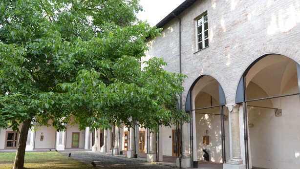 Ein Innenhof mit einem Gebäude, das von gewölbten Gangwegen gesäumt wird, und einem Baum, der Schatten spendet.