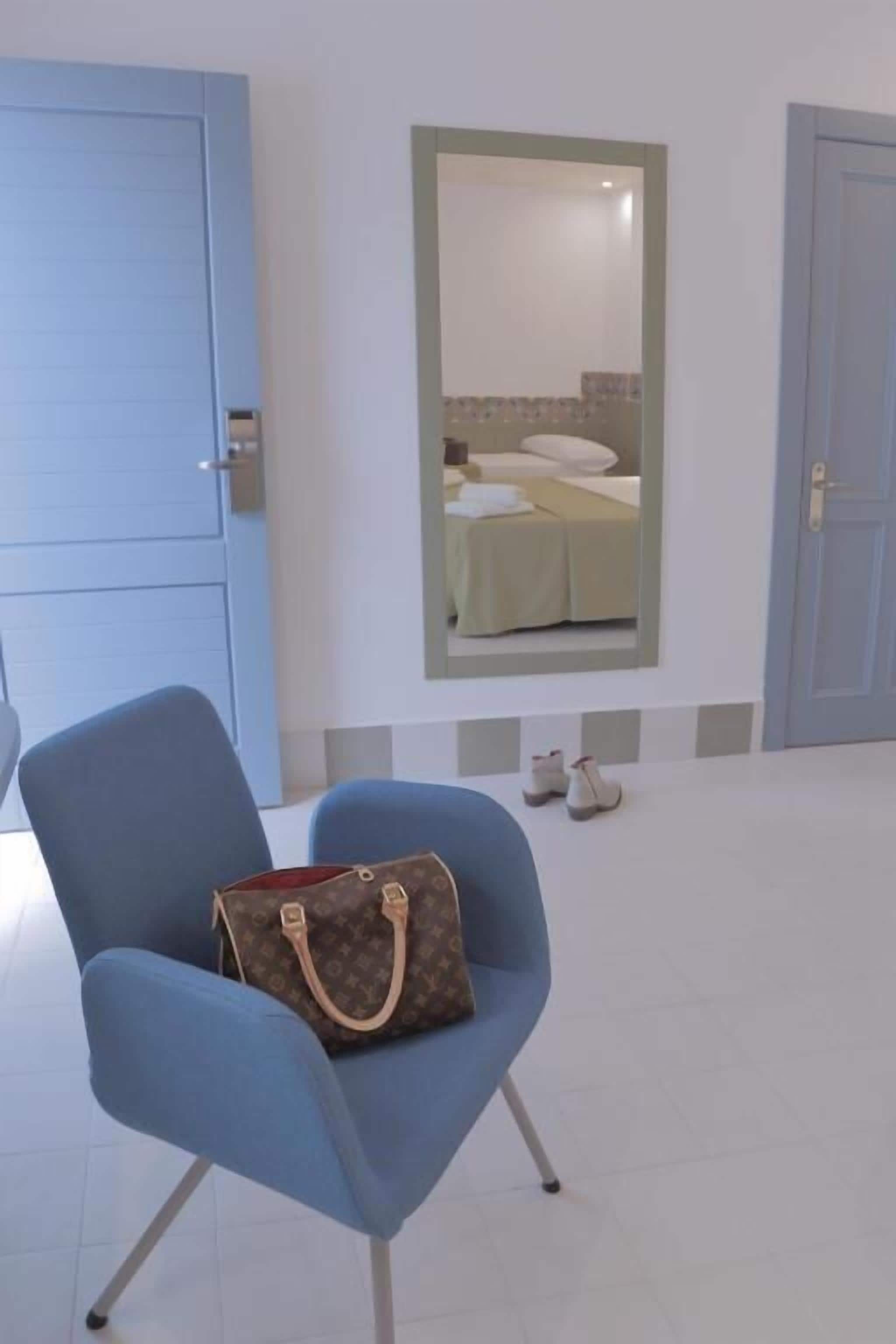Una habitación con un sillón azul, un bolso Louis Vuitton sobre él, un espejo en la pared y una cama con una colcha blanca.