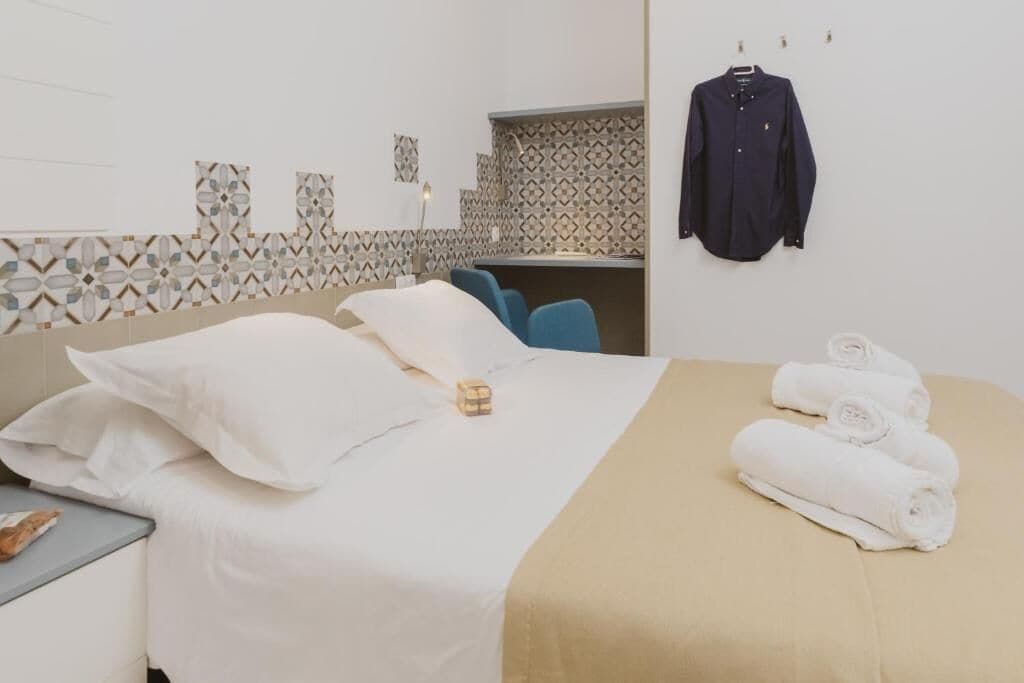 Une chambre d’hôtel avec un lit bien fait, une table de chevet avec un livre, un porte-serviettes avec des serviettes et un mur orné d’un motif.