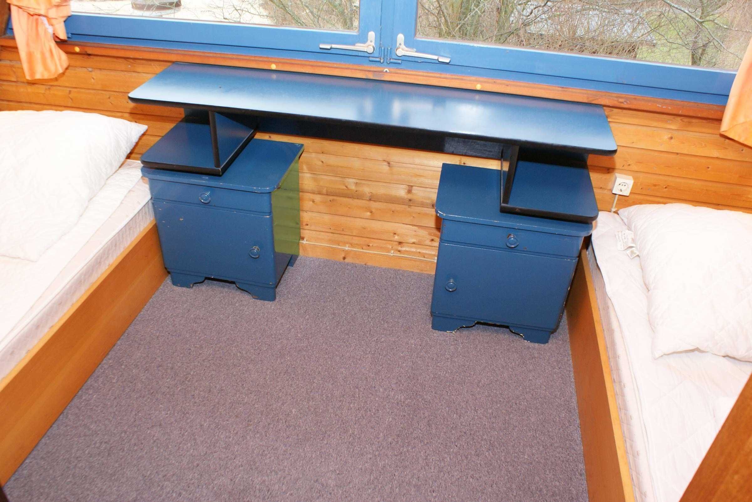 Une pièce de taille modeste comprenant un lit, deux tables de chevet bleues, un petit bureau et une fenêtre donnant sur des arbres.