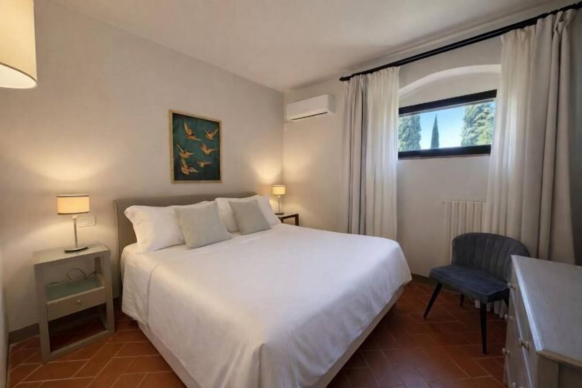 A hotel room with a bed, bedside lamps, a chair, a window with curtains, and an air conditioning unit.