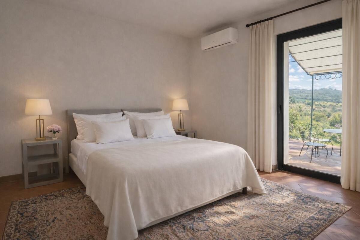 A bedroom with a bed, two bedside tables with lamps, a window with curtains, and a view of the outdoors.