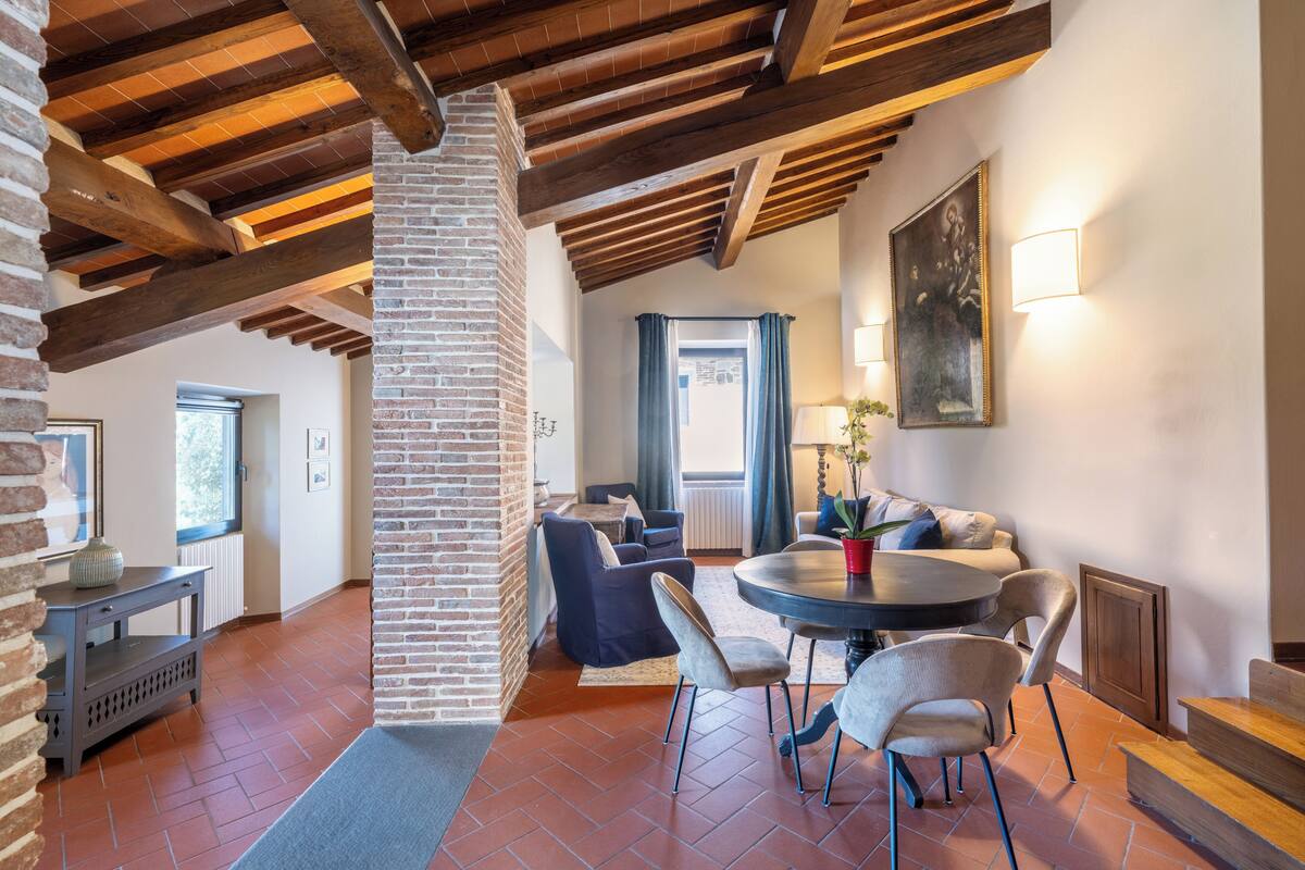 A spacious living area with a high ceiling, exposed wooden beams, a brick column, a round dining table with chairs, a sofa, and a window with curtains.