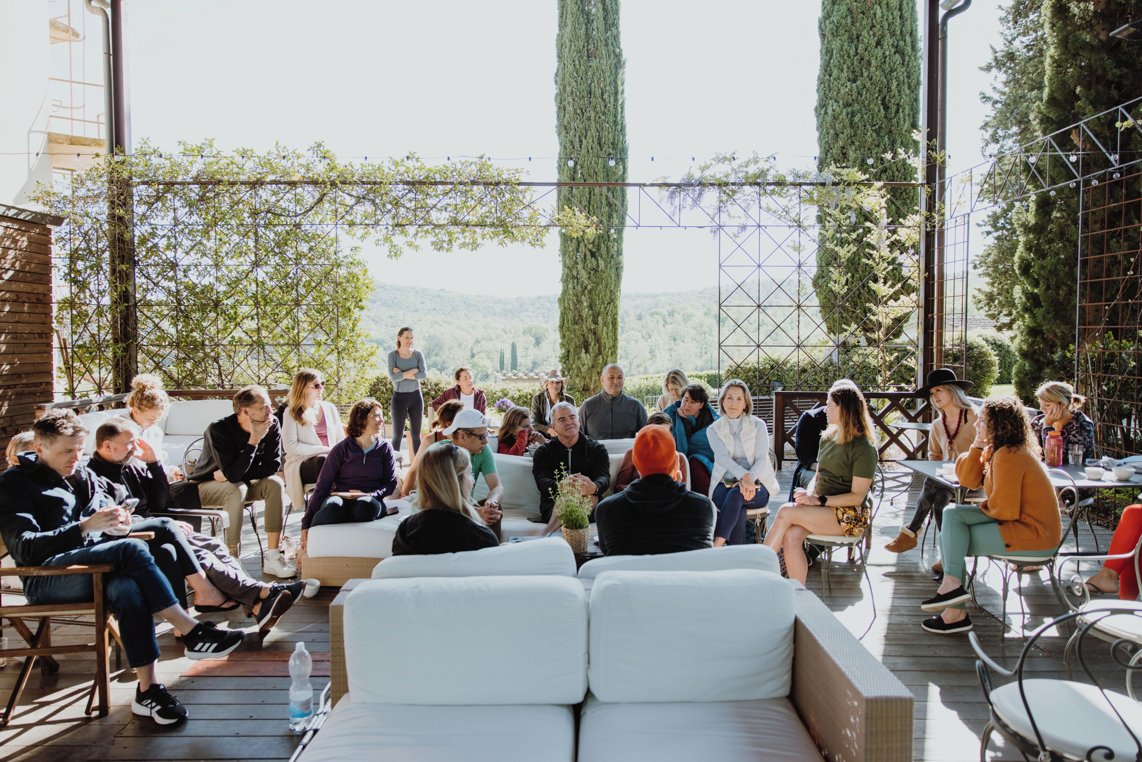 A group of people seated on outdoor furniture in a semi-open space with a view of trees and hills in the background.