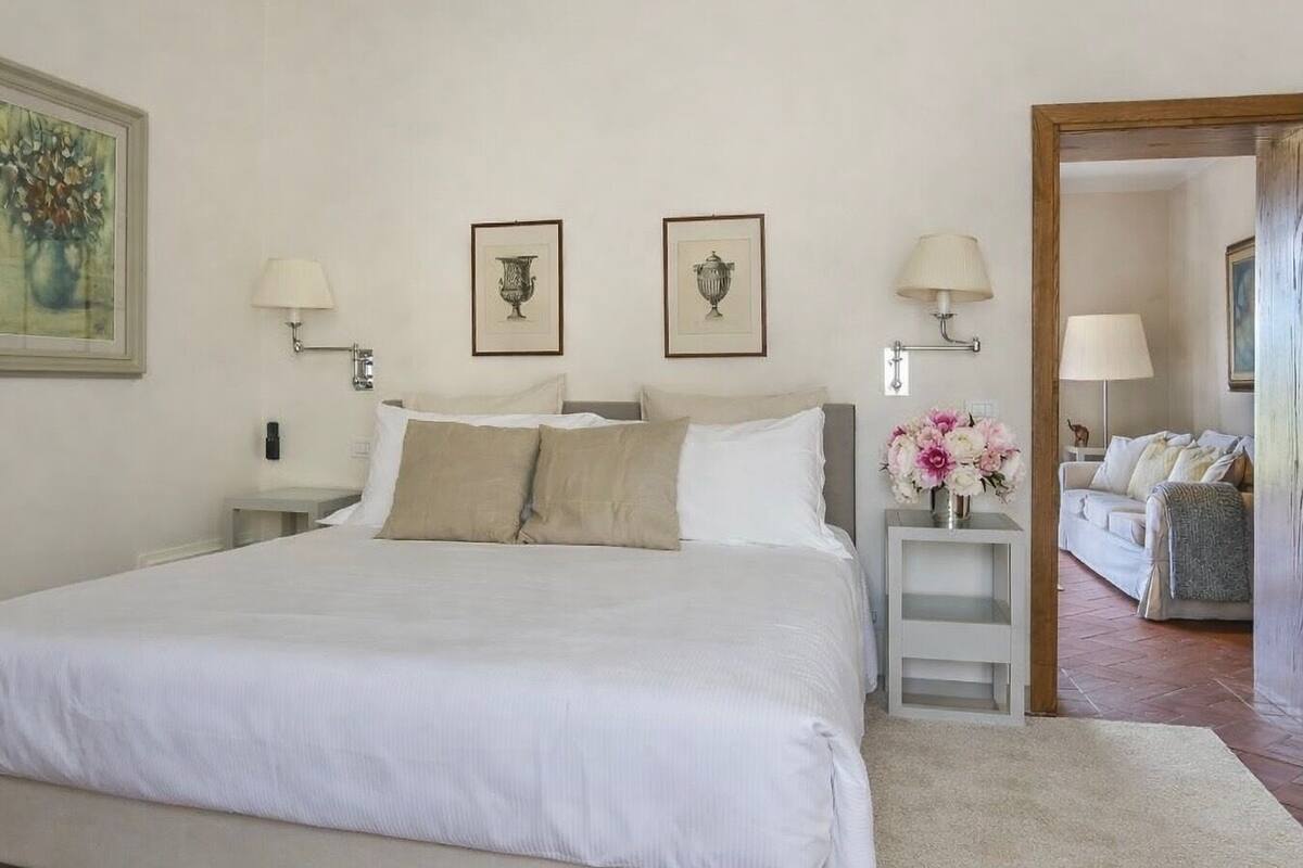 A neatly arranged bedroom with a large bed, two bedside tables, a nightstand, a small table with flowers, and a doorway leading to another room.
