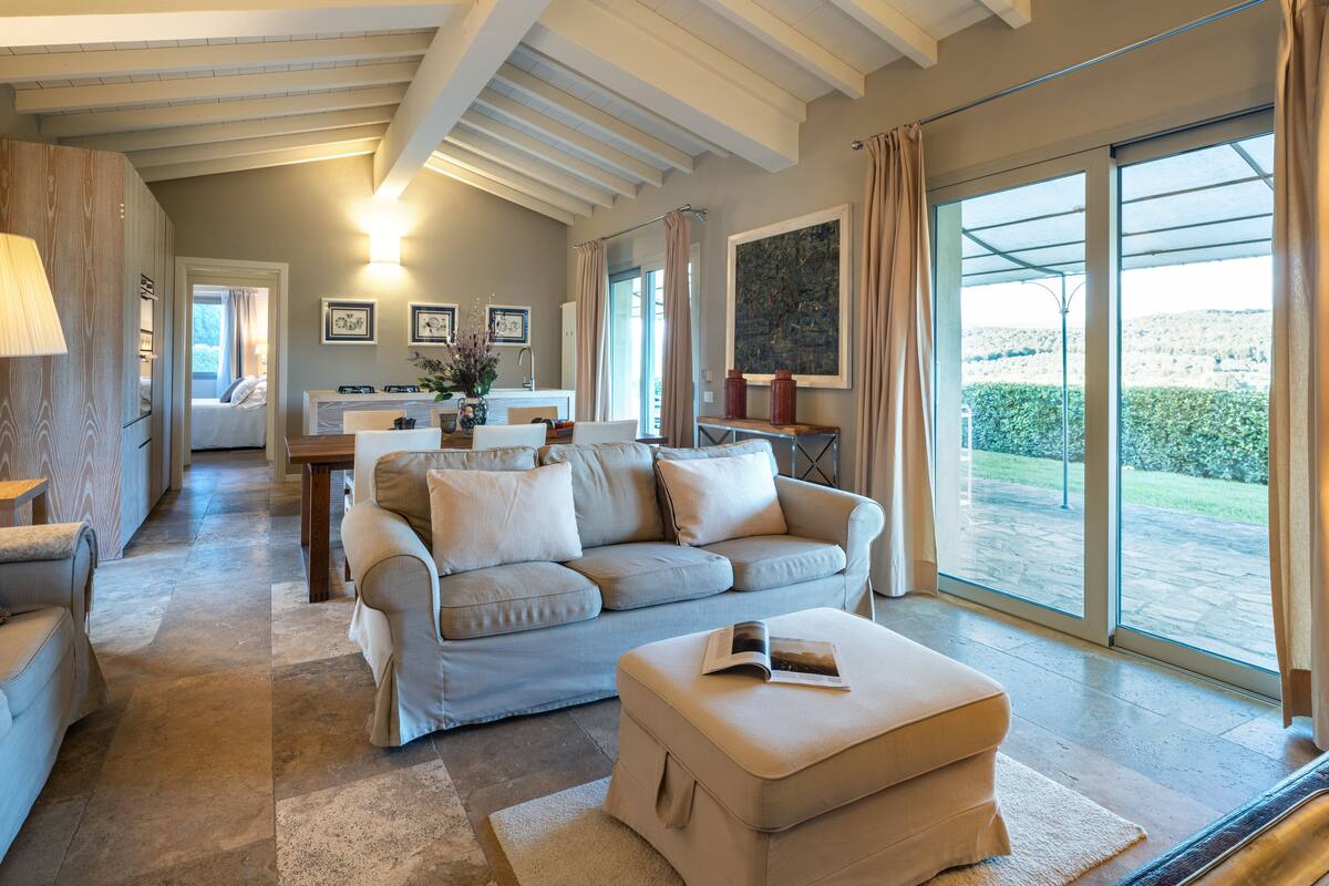 A spacious living area with a sofa, ottoman, and a dining table. A kitchen is visible in the background, and there is a large window offering a view of the outdoors.