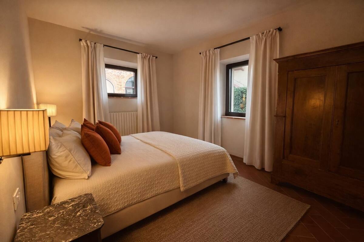 A bedroom with a bed, two windows with curtains, a wooden wardrobe, and a lamp.