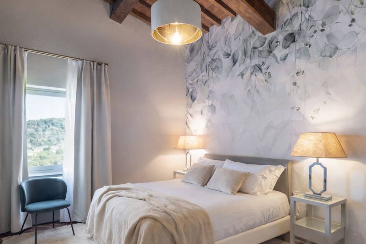 A bedroom with a bed, two bedside lamps, a chair, a large window with curtains, and a textured wall.