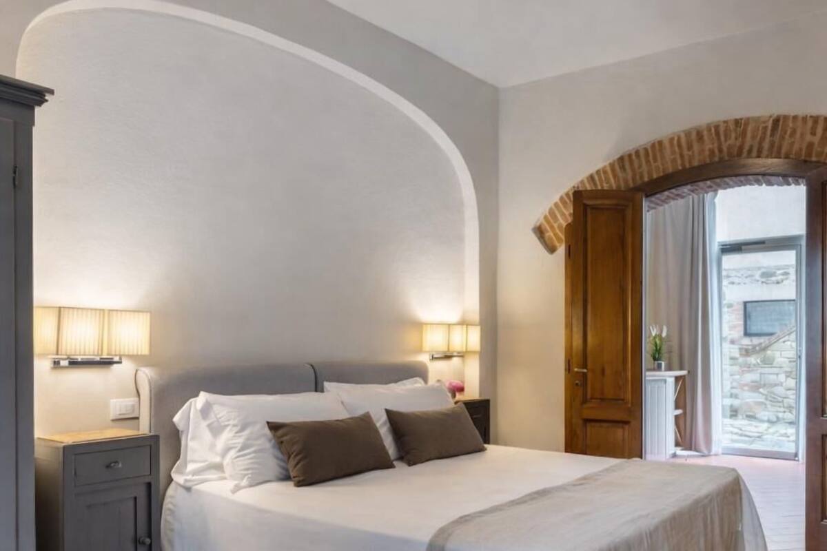 A bedroom with a large bed, two bedside tables with lamps, a wardrobe, and a view of a stone building through an arched doorway.