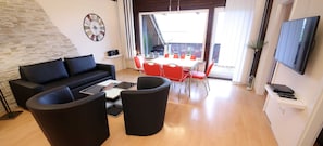 Un salon moderne avec un canapé, une table basse et des fauteuils rouges. On y trouve également une table à manger avec des chaises, un téléviseur fixé au mur et une horloge au mur.