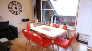 Uma área de refeições com uma mesa branca, cadeiras vermelhas, um relógio na parede e uma janela com vista para o exterior.