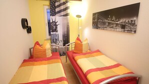 Quarto de hotel com duas camas, cada uma com roupa de cama e almofadas listradas. Há um telefone fixado na parede e um quadro com uma vista da cidade.