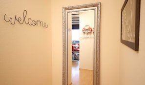 Un couloir orné d’une pancarte « Bienvenue », un miroir avec un cadre travaillé, et un aperçu d’un coin repas avec des chaises rouges et une table.